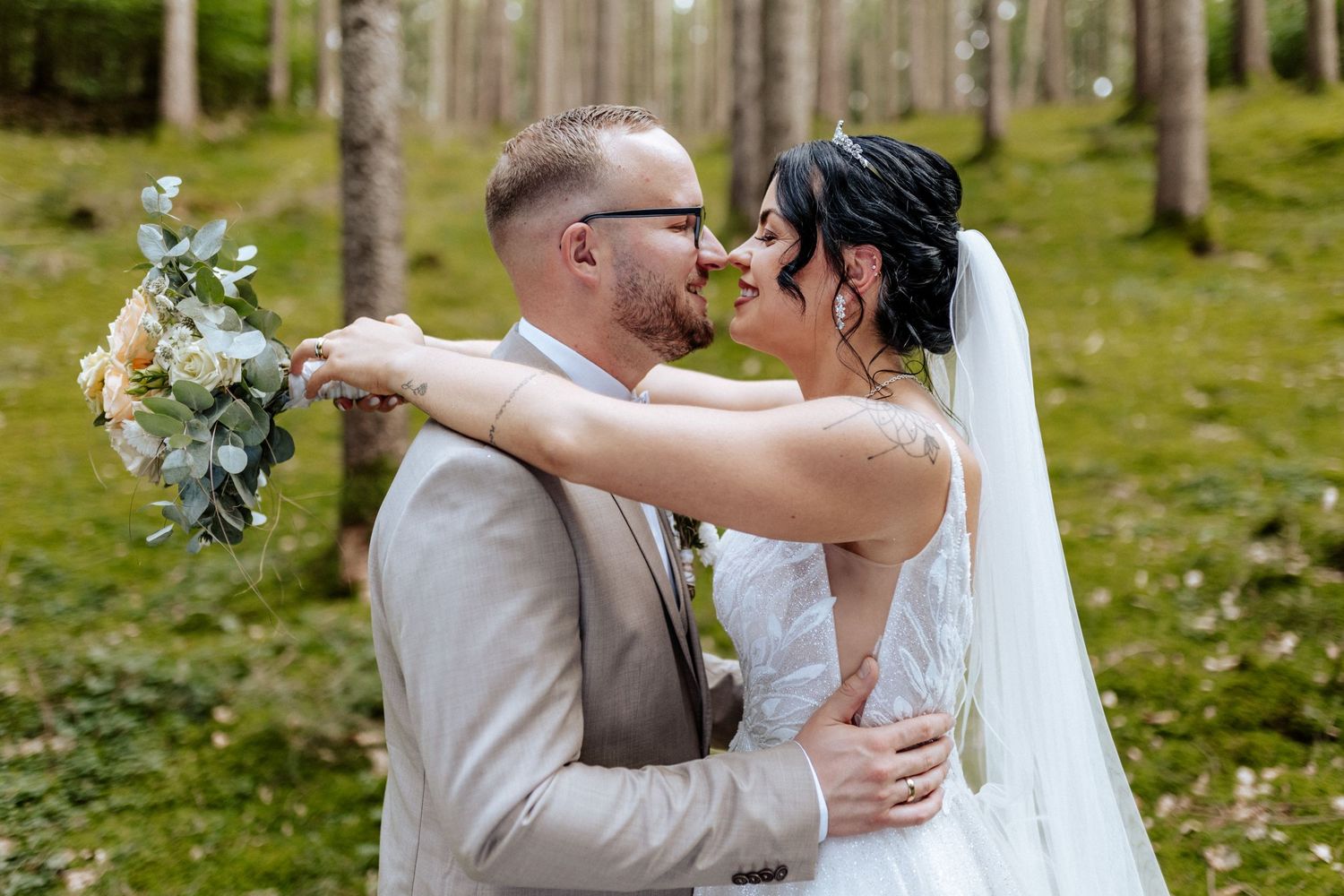 Couple sharing intimate moment in forest setting while bride holds floral bouquet.