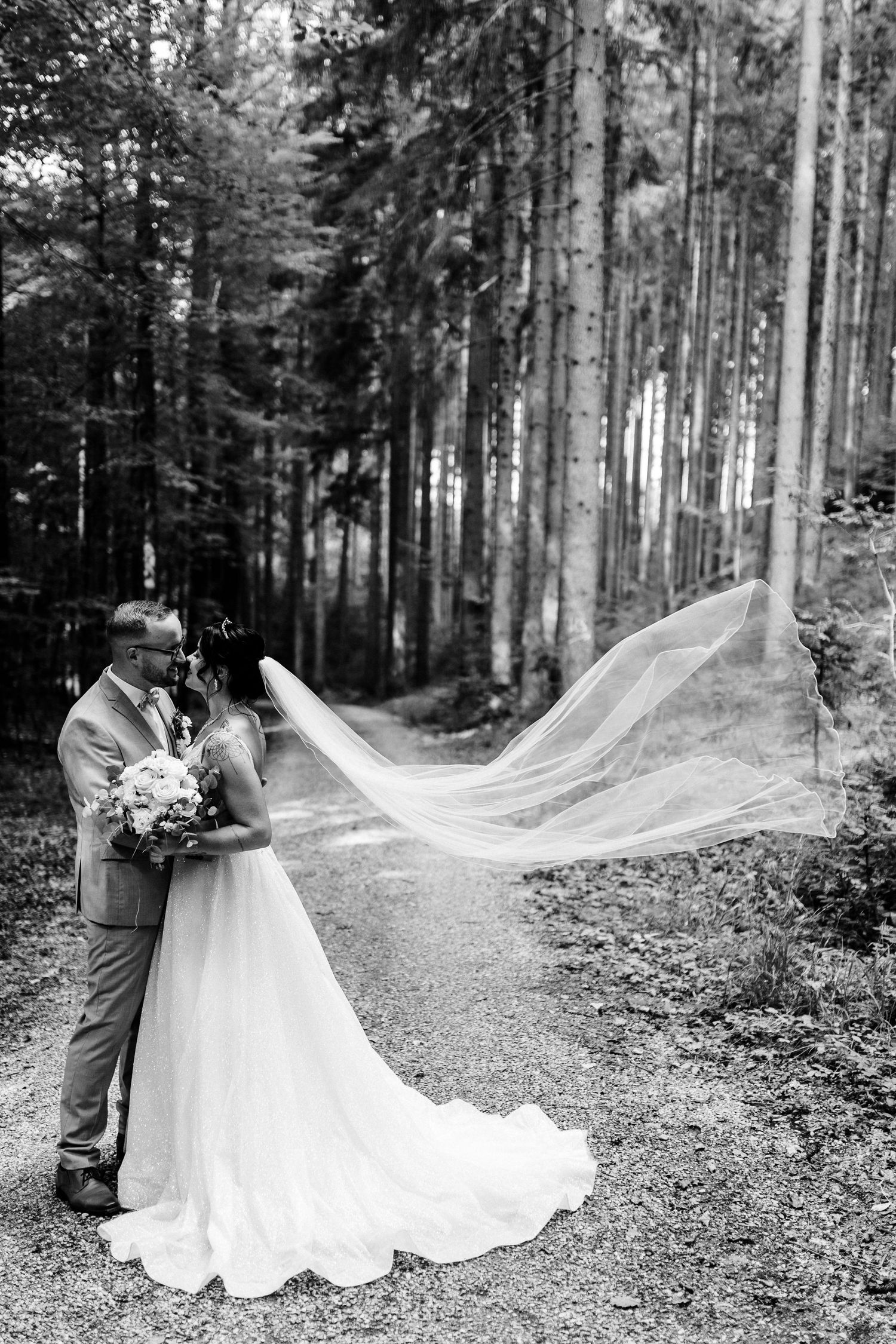 Romantic black and white portrait of couple embracing in forest with flowing veil caught in breeze.
