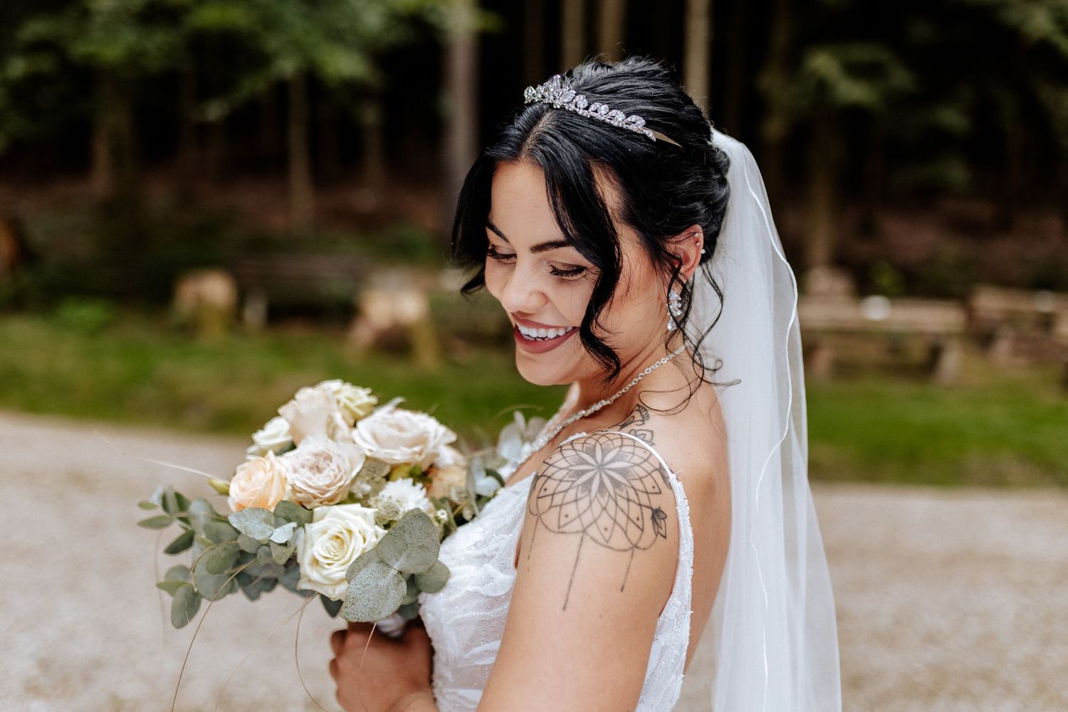 Bride holding white rose bouquet smiles while wearing delicate lace dress and sparkling hair accessory.