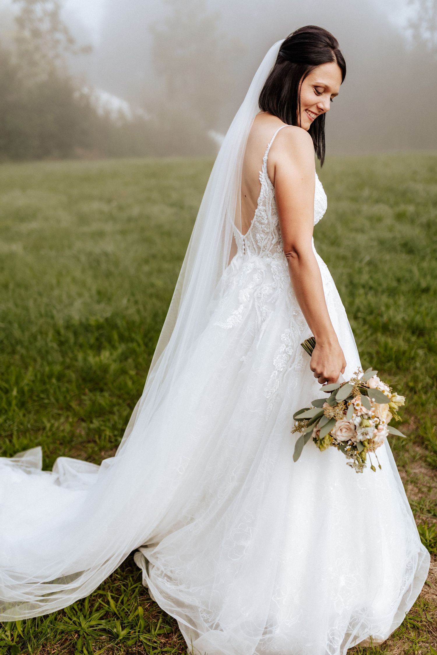 Bride in flowing white dress holding bouquet poses in misty field with long veil trailing behind.