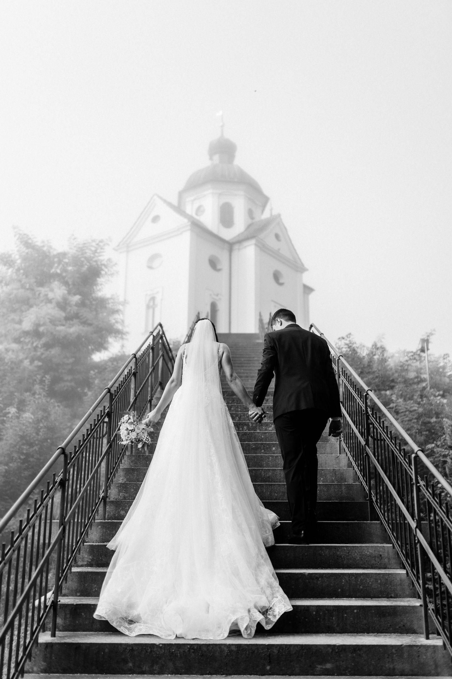 Newlyweds walking up stone steps hand in hand towards a white chapel in black and white.