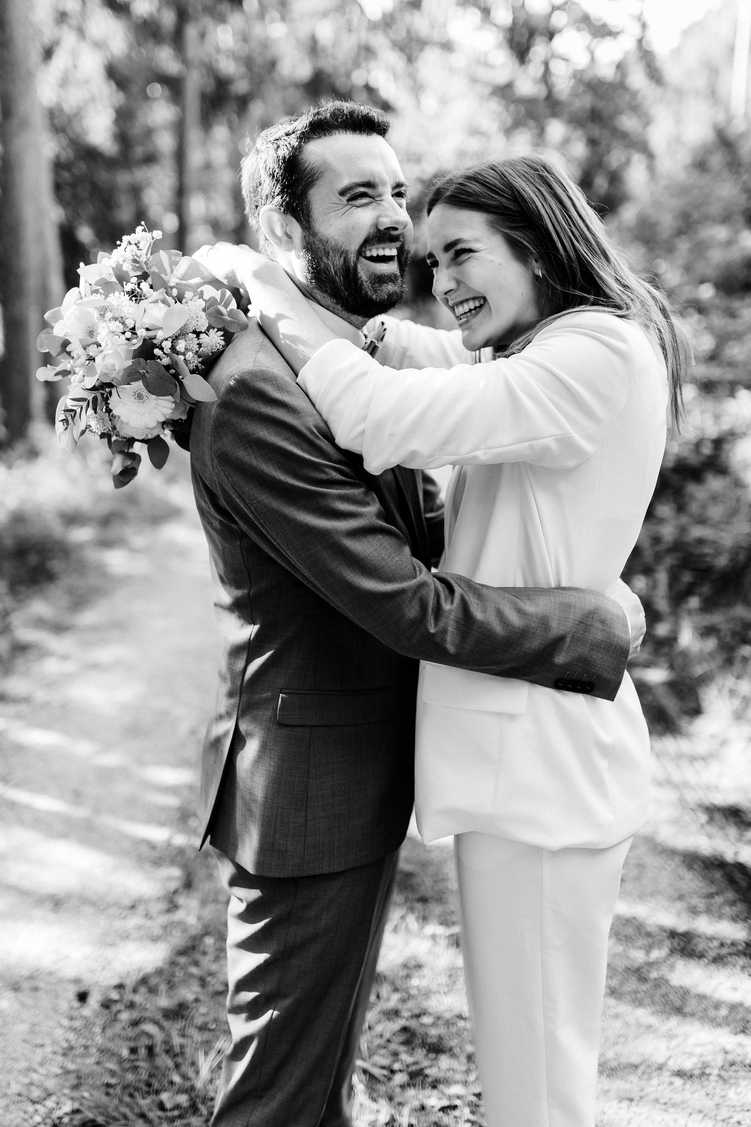 Happy couple embracing and laughing together while holding a floral bouquet in an outdoor setting.