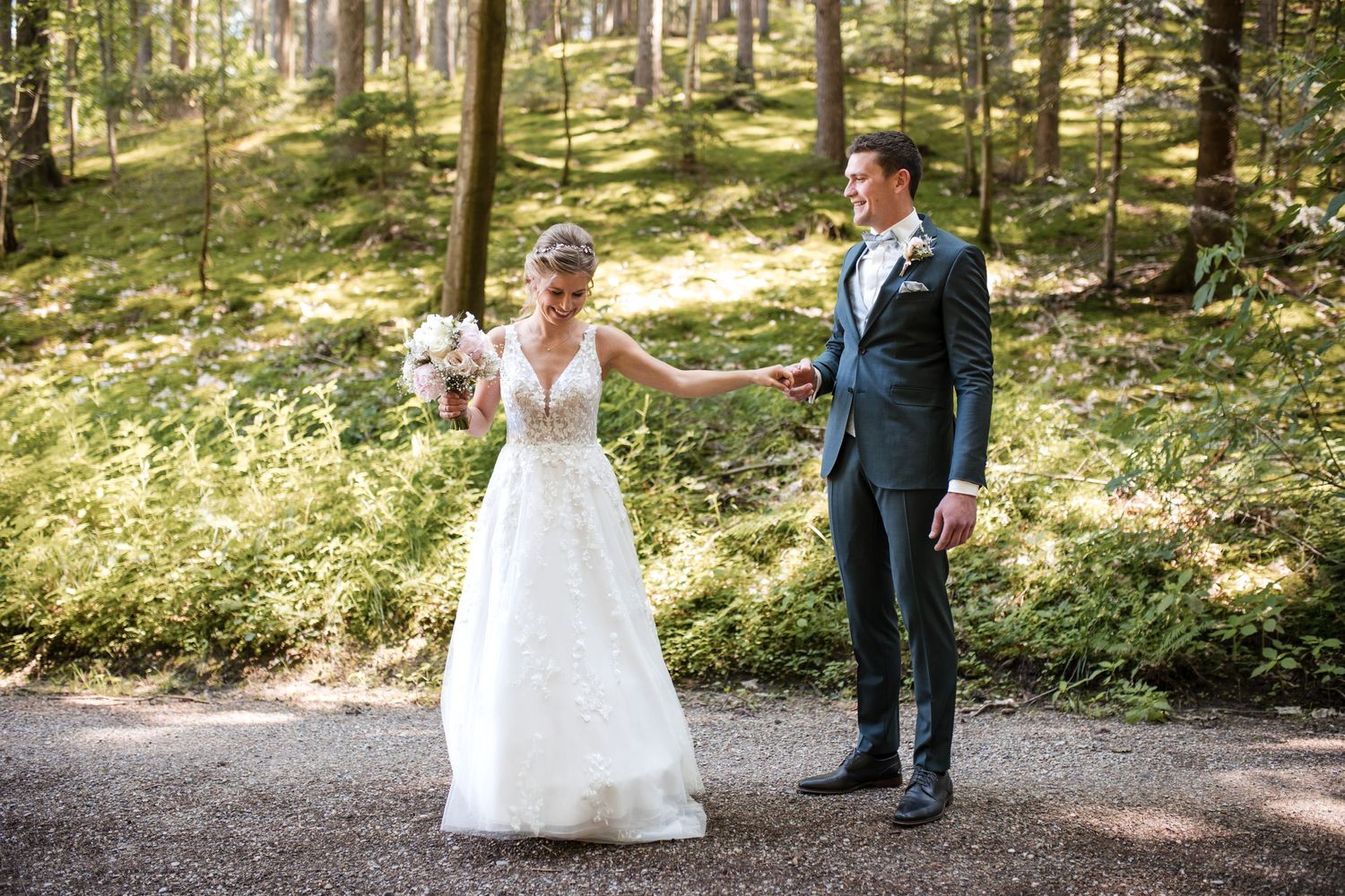 Newlyweds share a moment on a forest path, bride in lace gown and groom in navy suit.