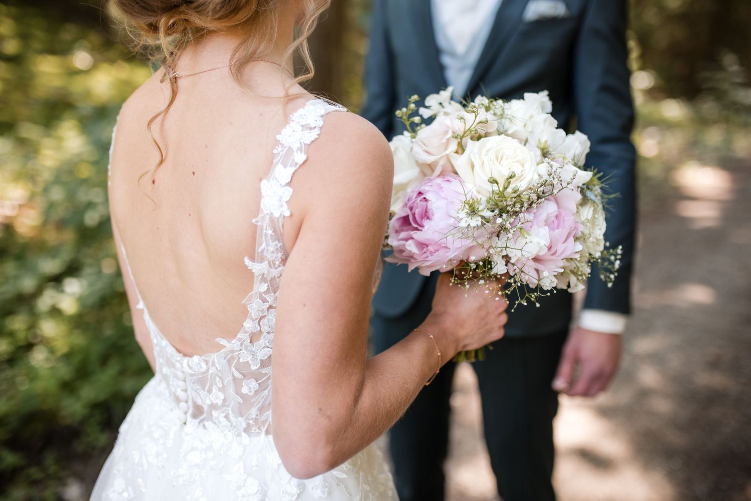 Detail shot of lace wedding dress back and pink and white floral bouquet against green outdoor backdrop.