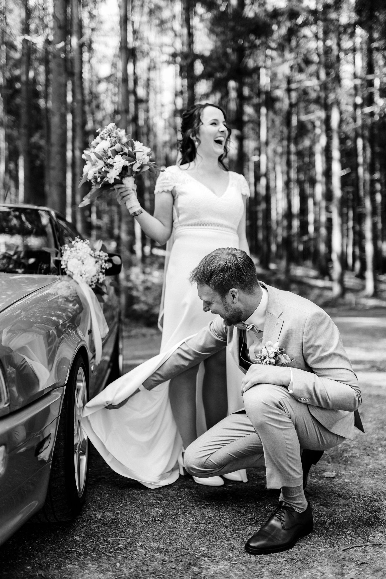 Candid moment with wedding dress and vintage car in forest setting.