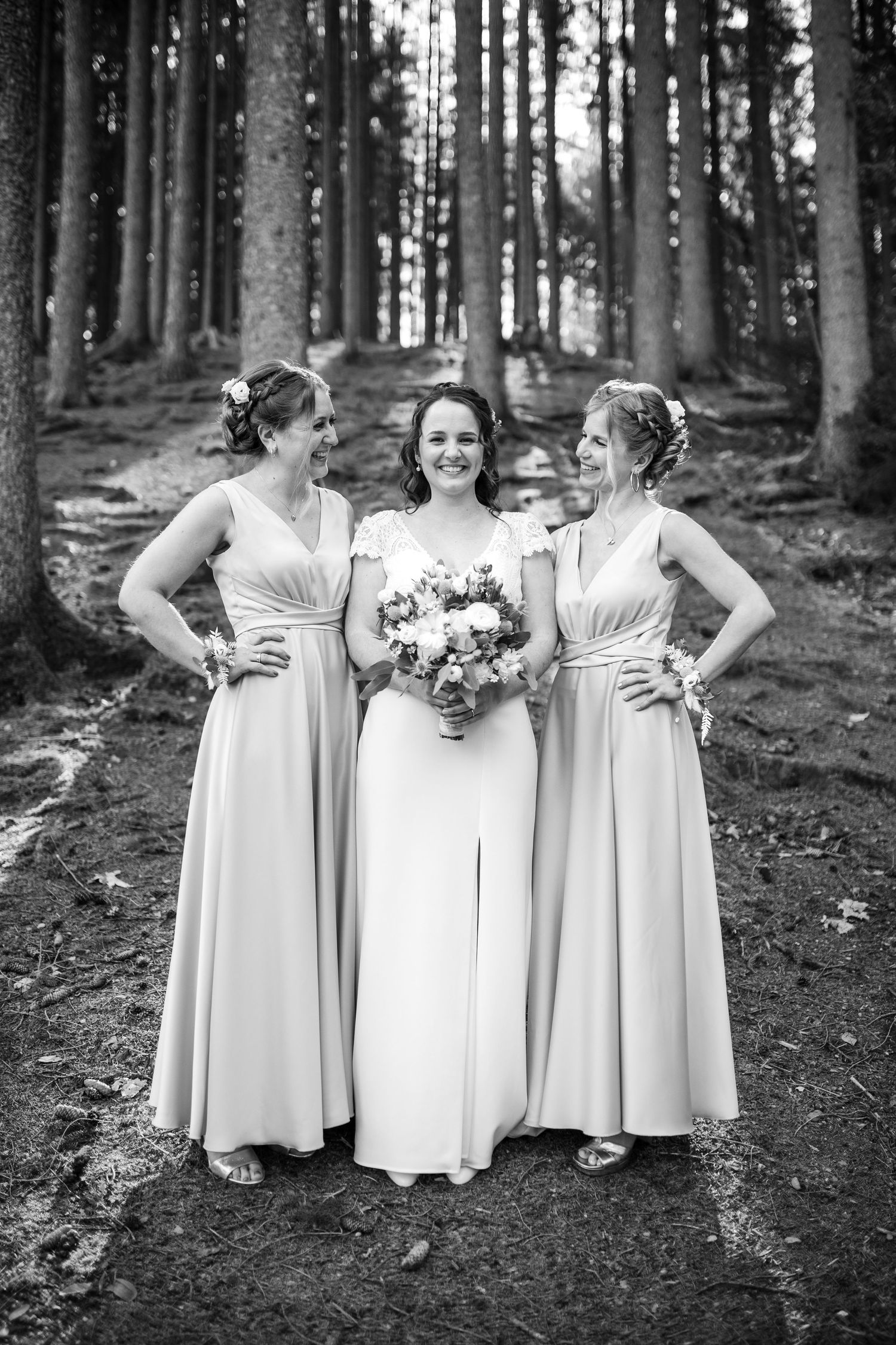 Three women in formal dresses pose together in a forest setting.
