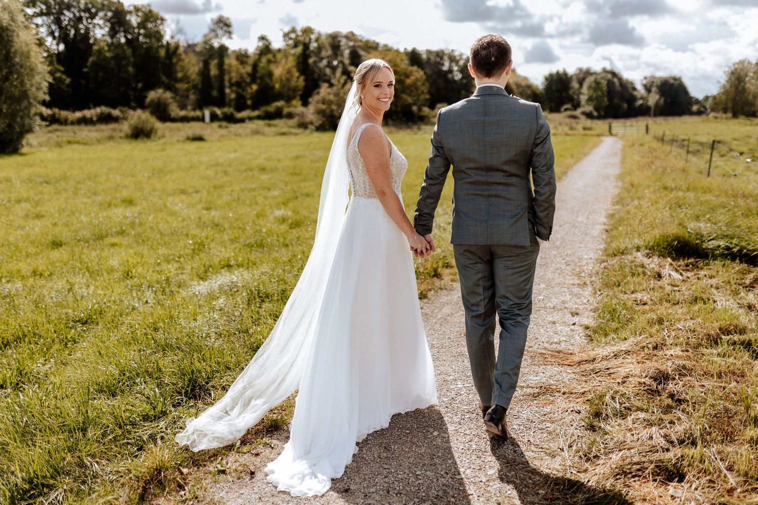 Newlyweds walking hand in hand down a country path with flowing veil.