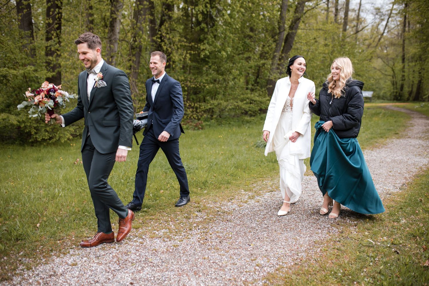 Wedding party walks along a gravel path through lush forest with bridesmaids in formal attire and bouquets.