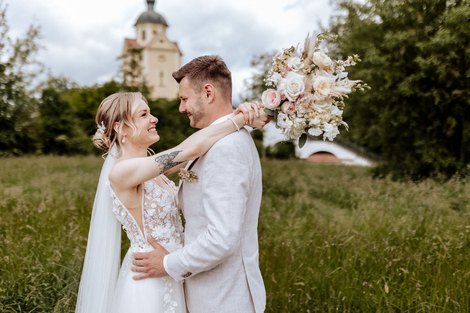 Couple in white wedding attire embracing in a sunny meadow with florals.