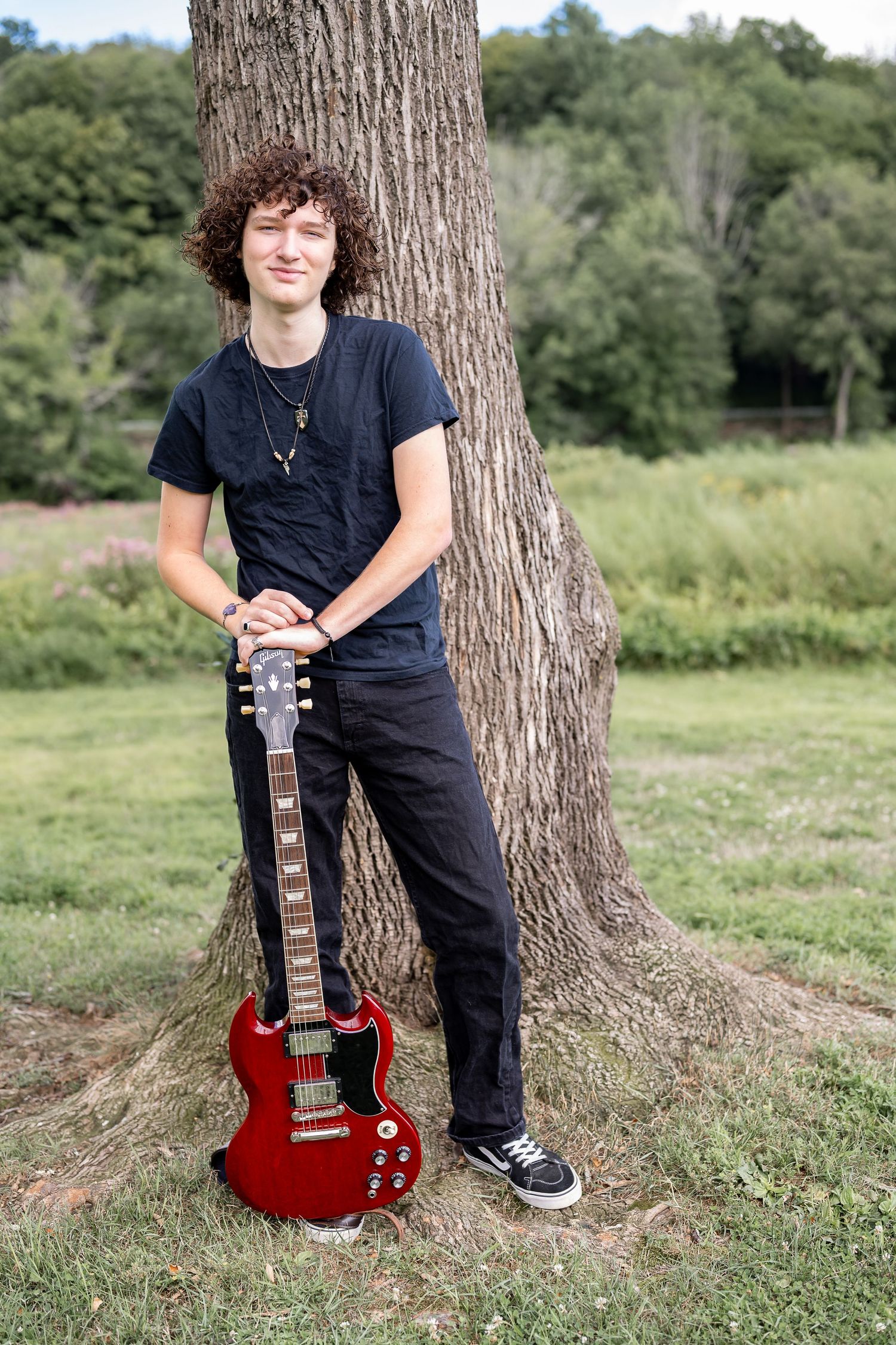Musician posing with red electric guitar against large tree trunk in outdoor setting for his senior session in Massachusetts.