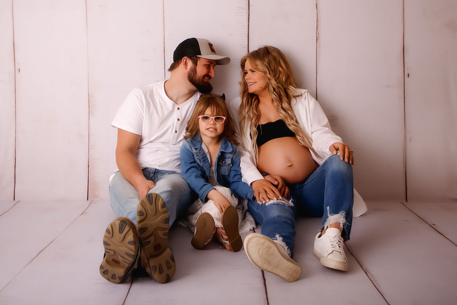 A casual family portrait against white wooden backdrop shows expecting mother with loved ones.
