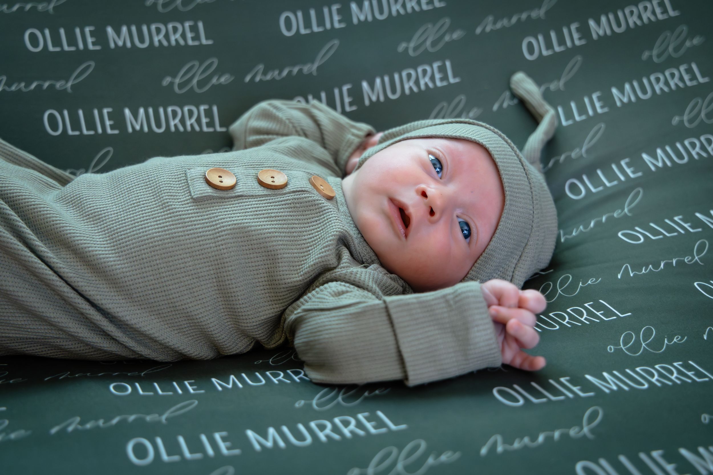 A newborn baby wrapped in beige swaddle blanket lies on a green chalkboard with repeating text.