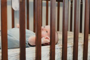 A small baby's feet peek through dark wooden crib slats against a light-colored background.