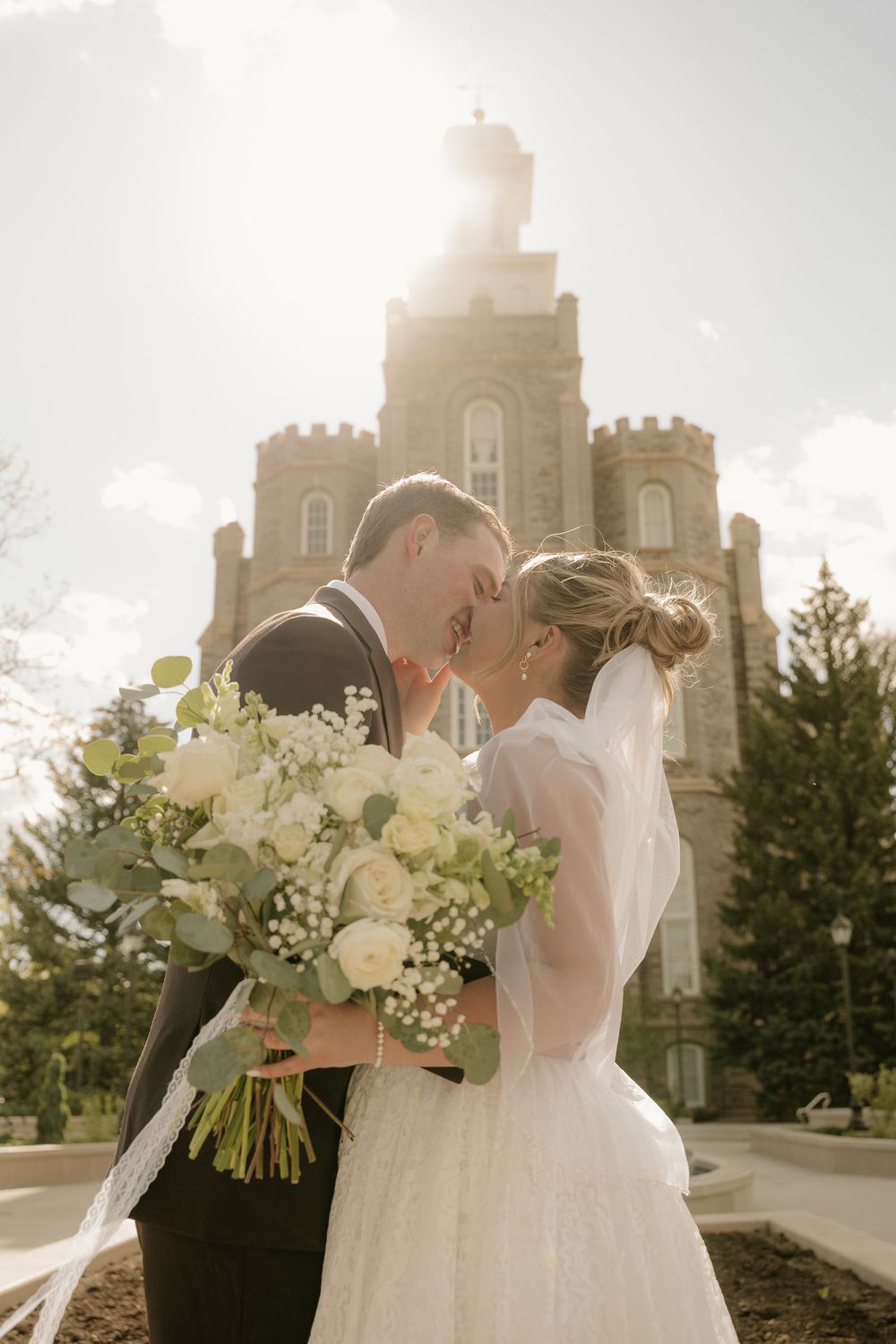 Bride and groom share a romantic moment holding white floral bouquet in front of a sunlit stone castle.