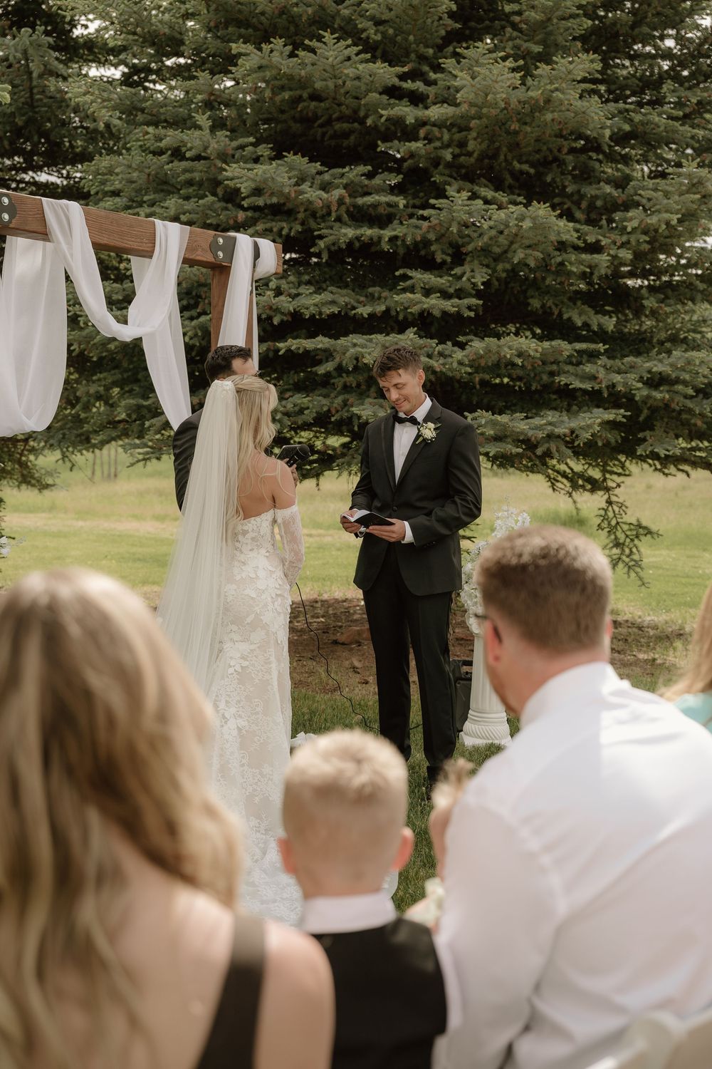 Outdoor wedding ceremony with groom in black tux reading vows to bride in lace dress under wooden arch with white draping.
