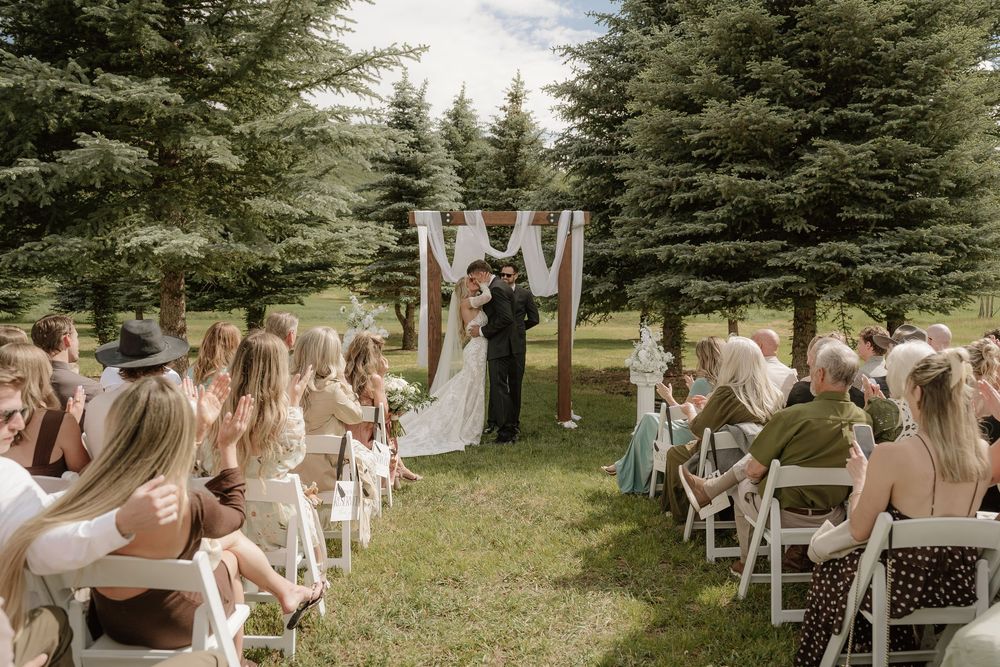 Outdoor wedding ceremony with couple kissing at floral arch altar, guests seated in white chairs on green lawn surrounded by pine trees.