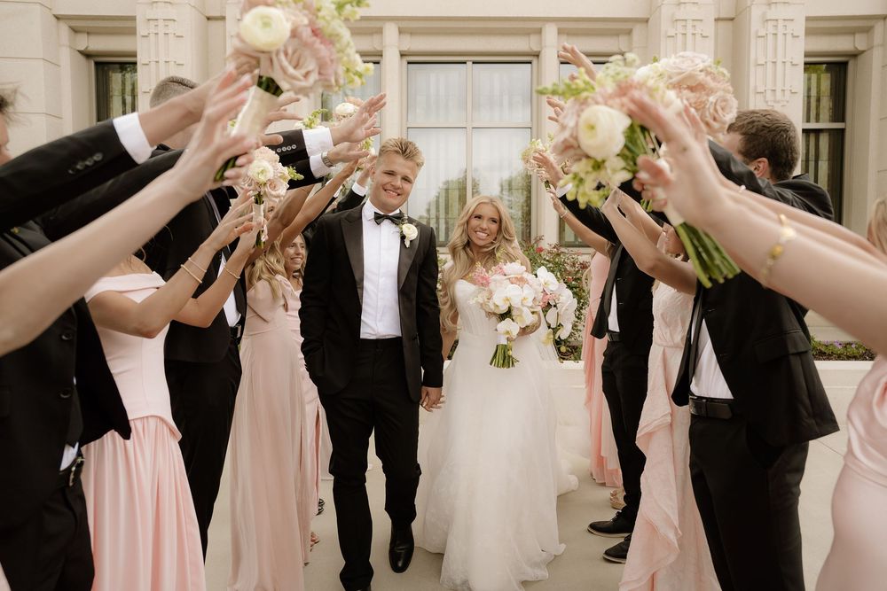 Bride and groom walk through an arch of raised bouquets with bridesmaids in pink and groomsmen in black tuxedos.