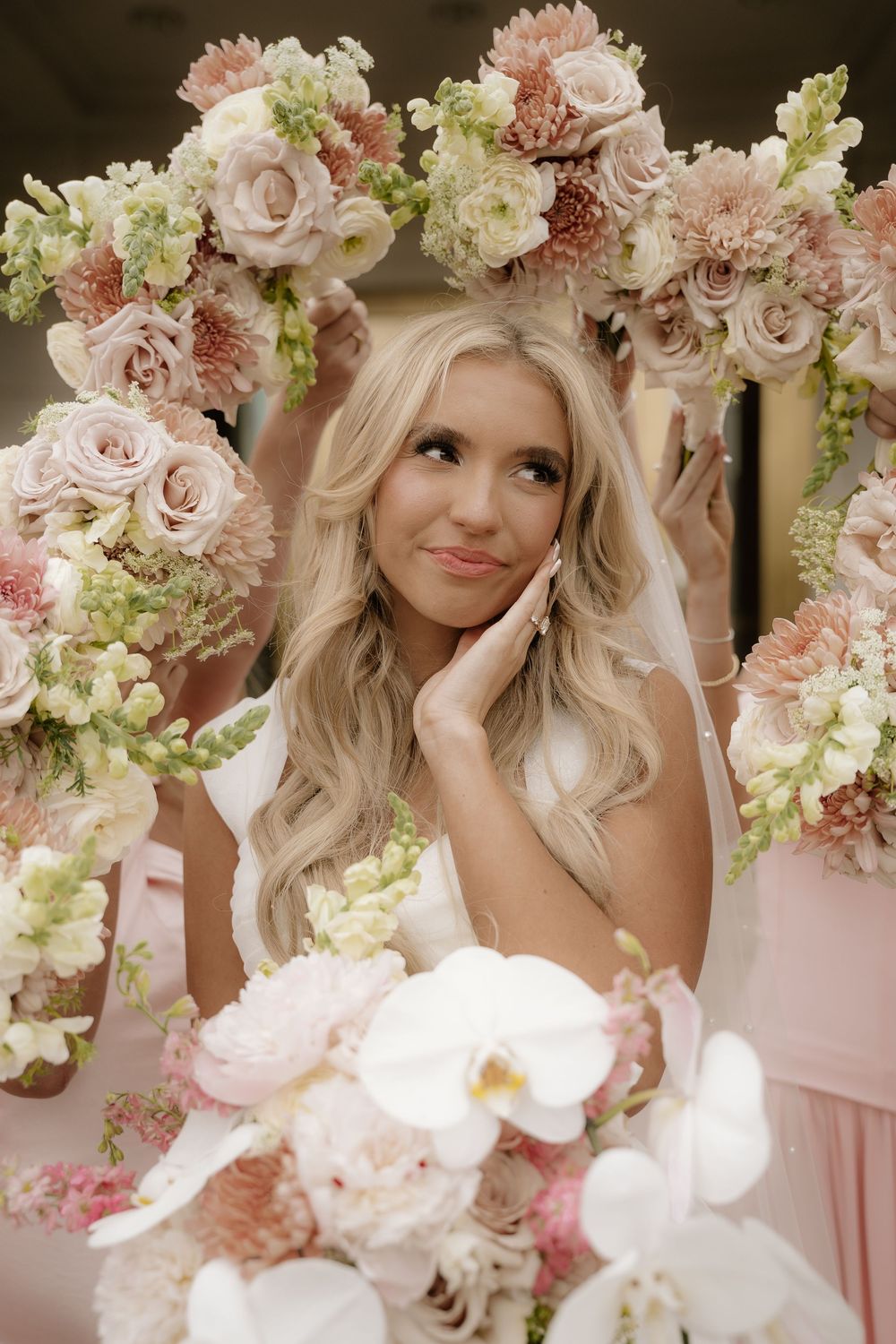 Bride surrounded by pink and white floral bouquets, posing thoughtfully in a mirror reflection with bridesmaids.