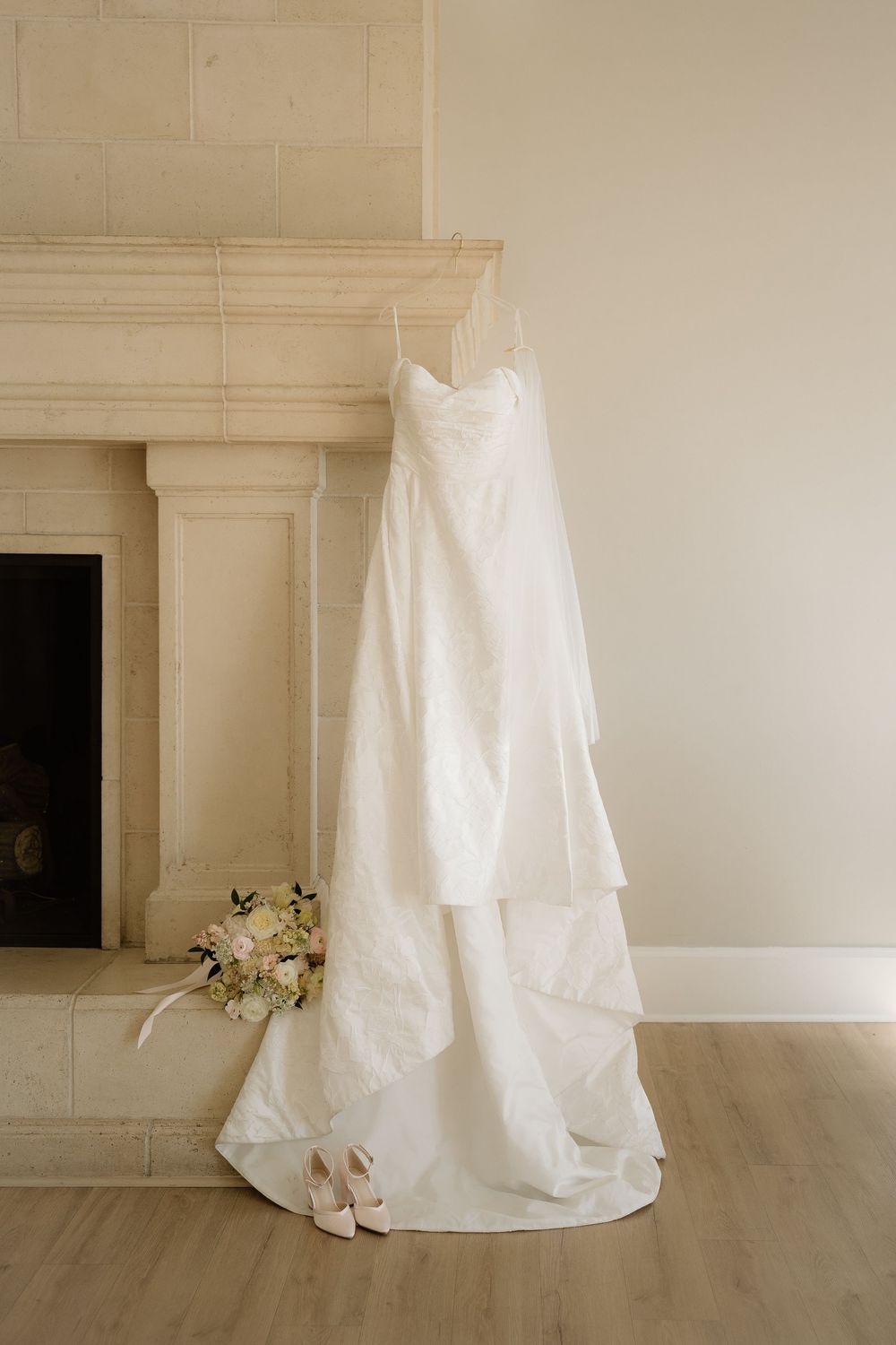 White wedding dress hanging above fireplace with bridal bouquet and heels on hardwood floor.