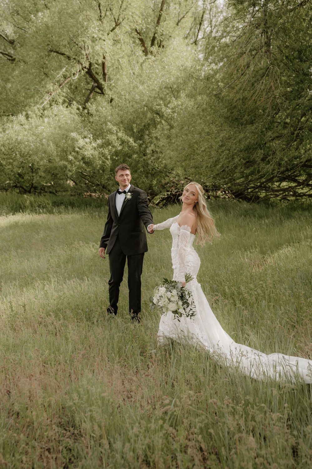 A bride and groom walk hand-in-hand through a lush green meadow, she wearing a lace gown with a long train.