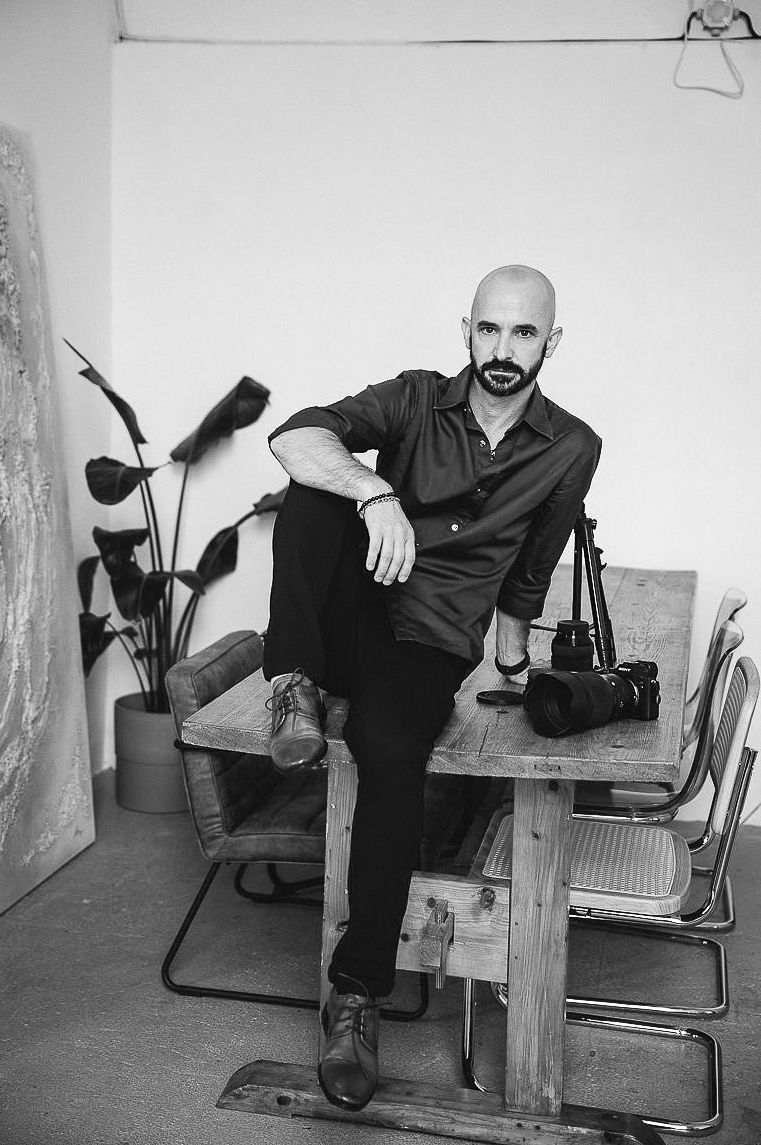 Black and white portrait of a bald bearded photographer sitting on a wooden table with camera equipment in a studio.