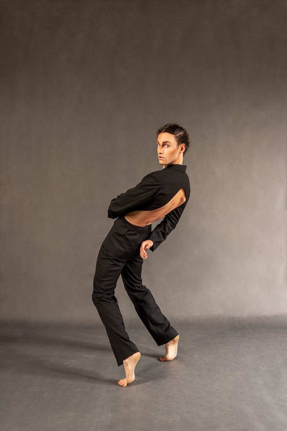 Contemporary dancer in black open-back outfit poses dynamically, looking over shoulder in studio setting.
