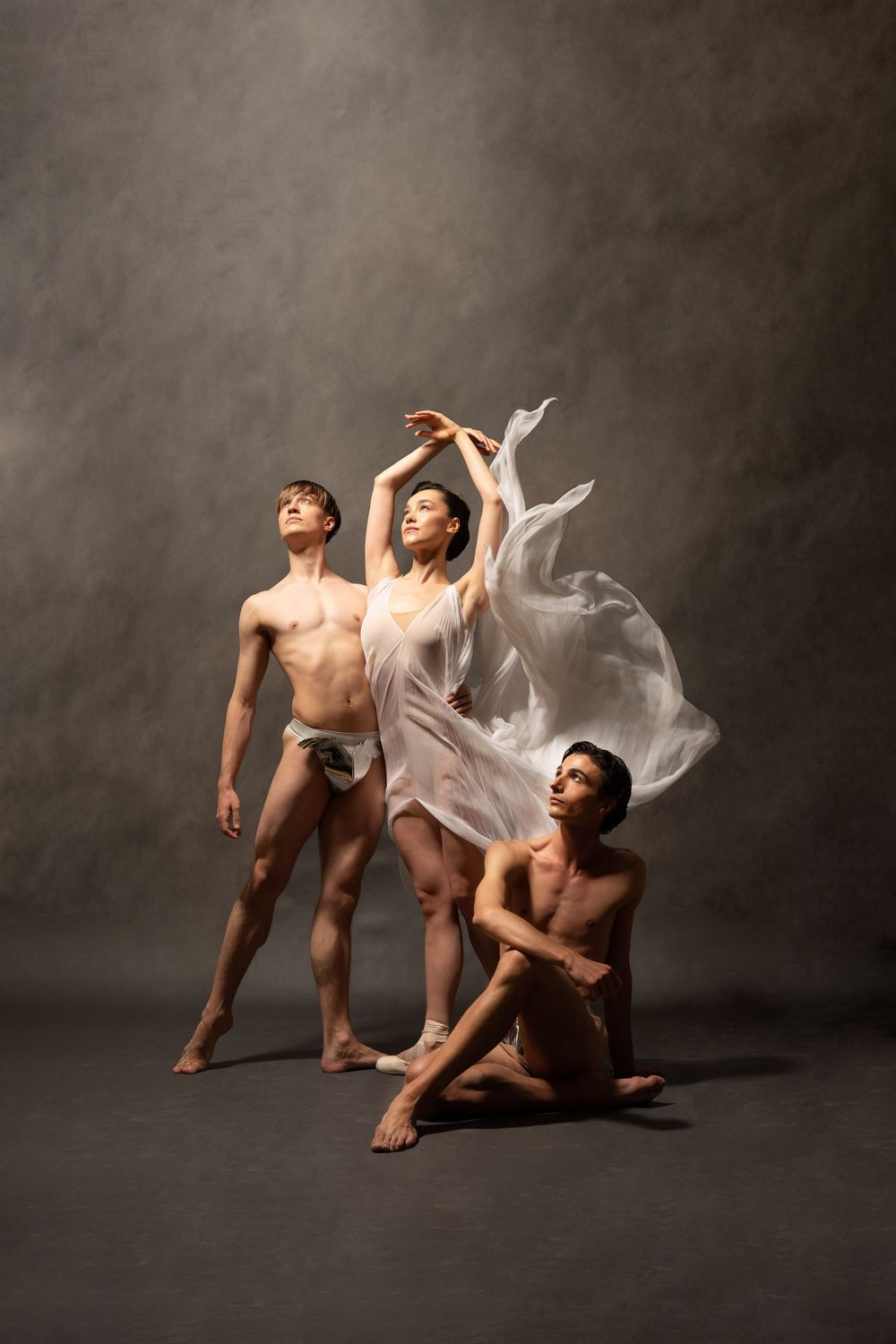 Three ballet dancers in white and minimal costumes perform elegant group pose with flowing fabric against gray wall.