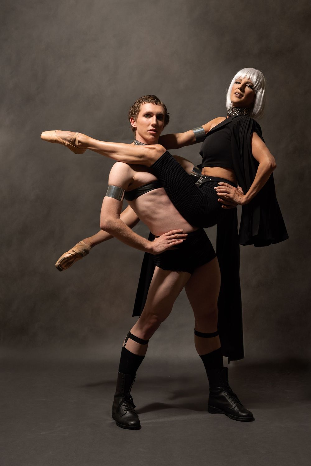 Male ballet dancer lifts leg while female partner in black supports him in studio.