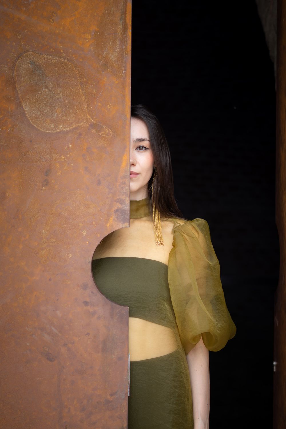 Woman in olive green and yellow organza dress peers around a weathered rusty metal panel into a dark doorway.