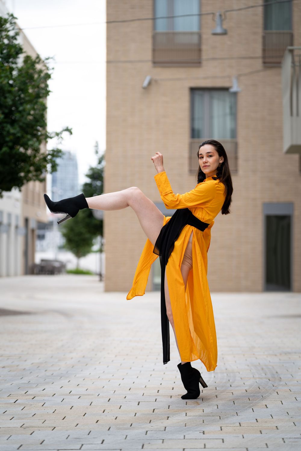 Female dancer in bright orange dress and black heels kicks her leg high on an urban city street outdoors.