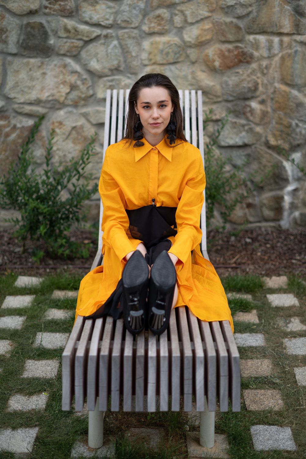 Woman in yellow shirt dress sits on wooden bench holding black heels against stone wall.