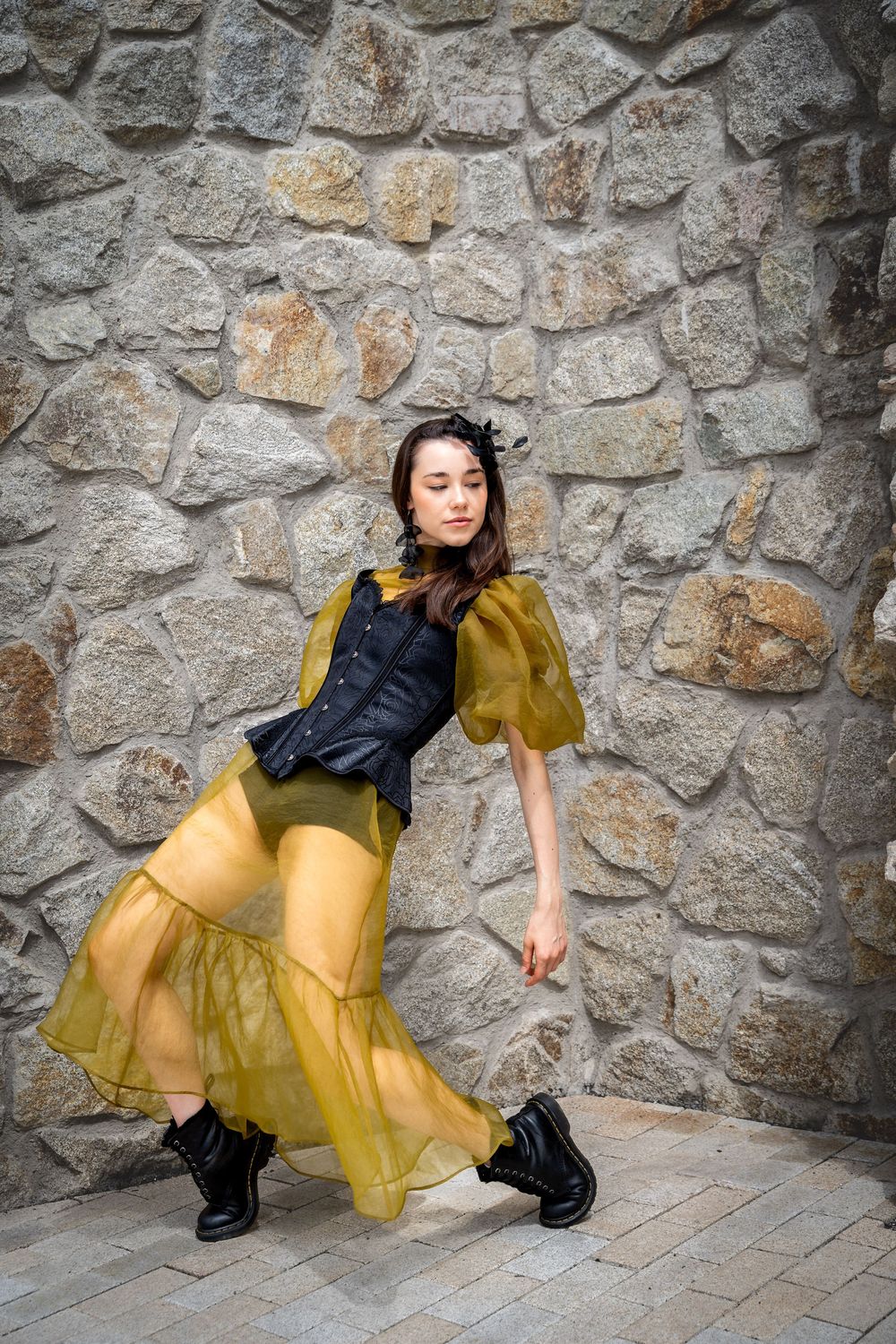 Female dancer in olive yellow organza dress and black boots poses gracefully against a rustic stone wall outdoors.
