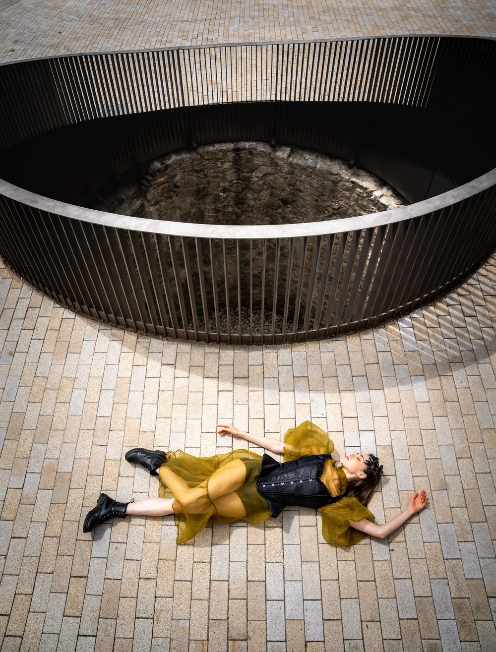 Female dancer in yellow organza dress and black boots lies dramatically on decorative tiled pavement near a circular fountain.