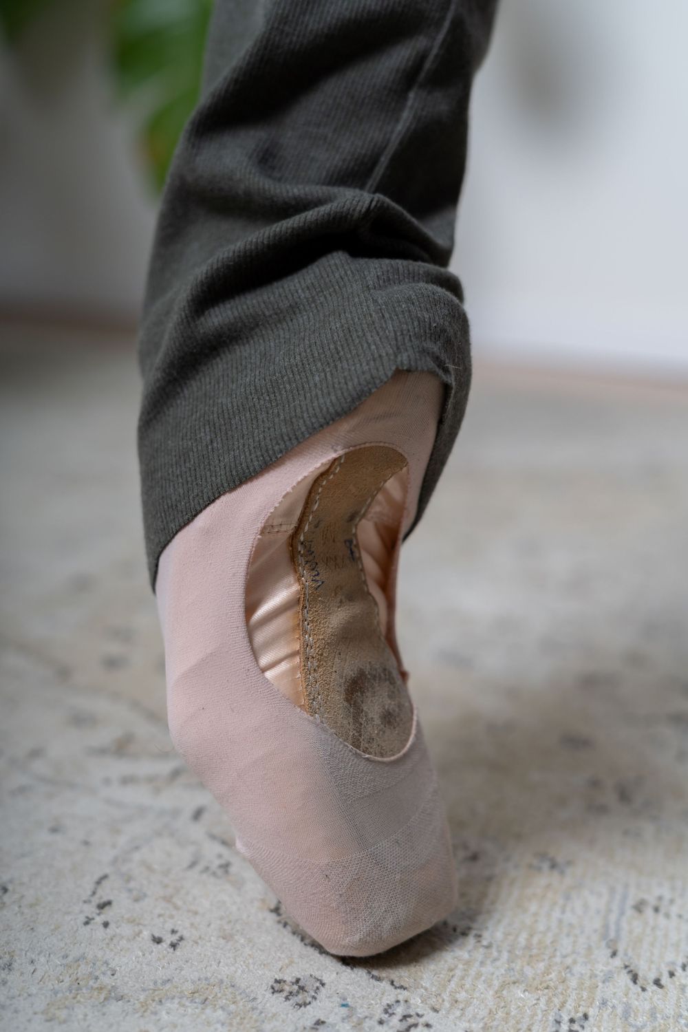 Close-up of ballet dancer foot in worn pink pointe shoe on gray carpet floor.