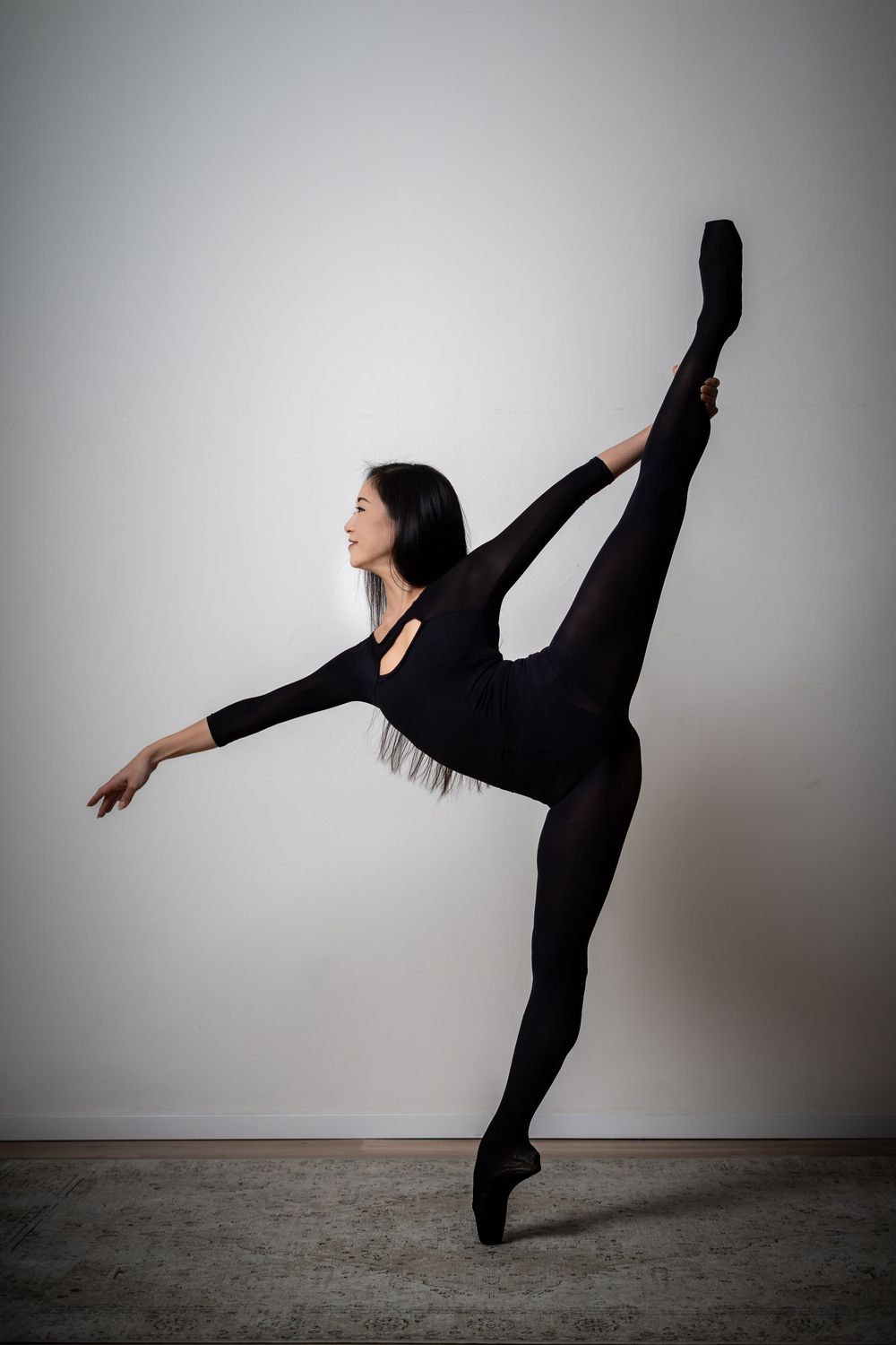 Ballet dancer in black leotard performs high leg extension against white studio wall.