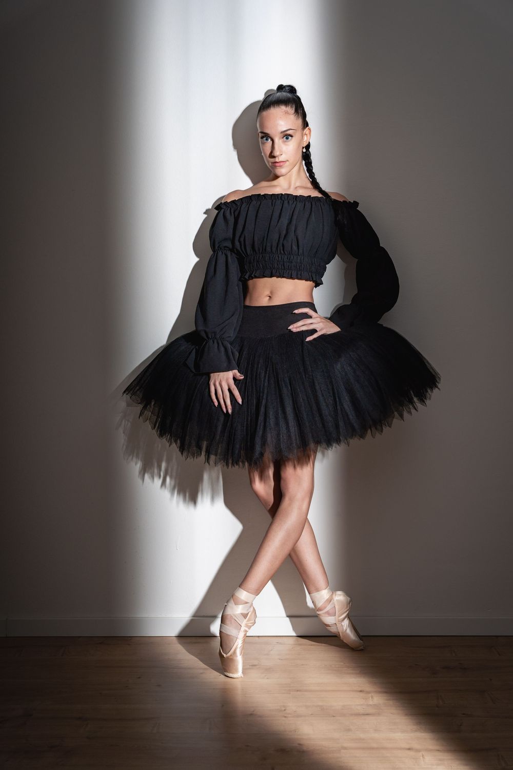 Ballerina in black ruffled crop top and tutu stands in pointe shoes against gray wall.