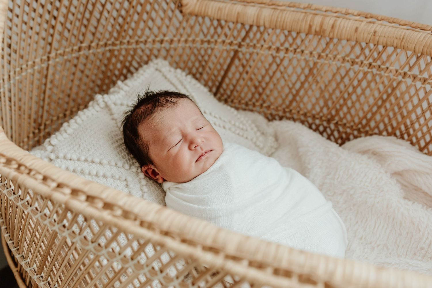 A sleeping newborn baby swaddled in white blanket rests in a woven basket.