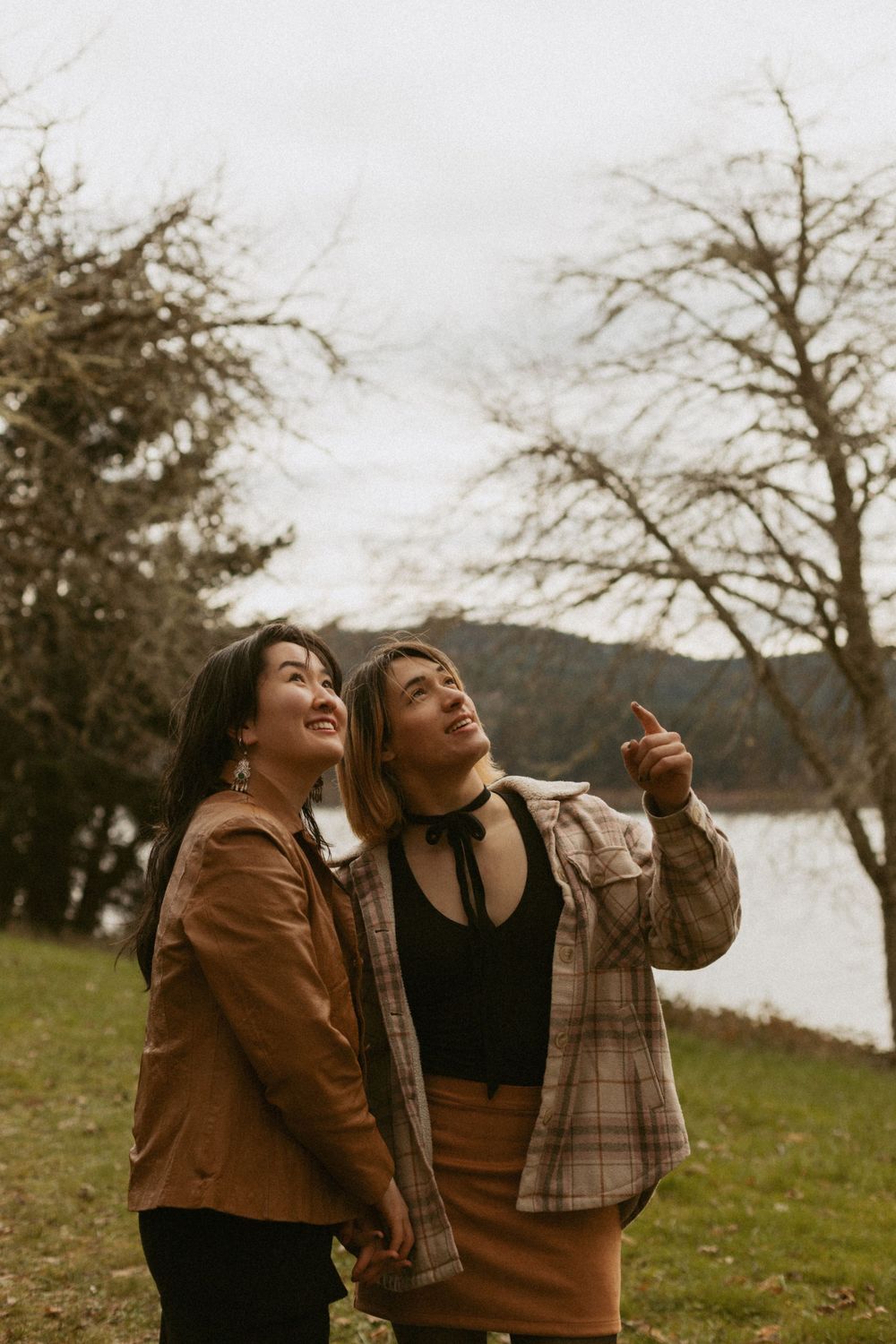 Two people in casual wear laughing and pointing at something in the distance by a lakeside.