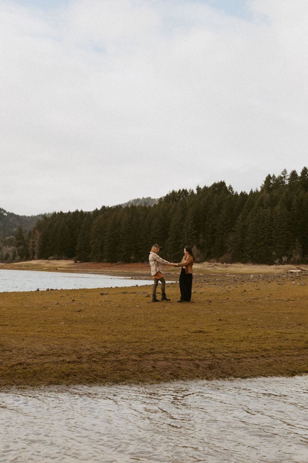 People stand on sandy shoreline with evergreen forest and mountains in background.