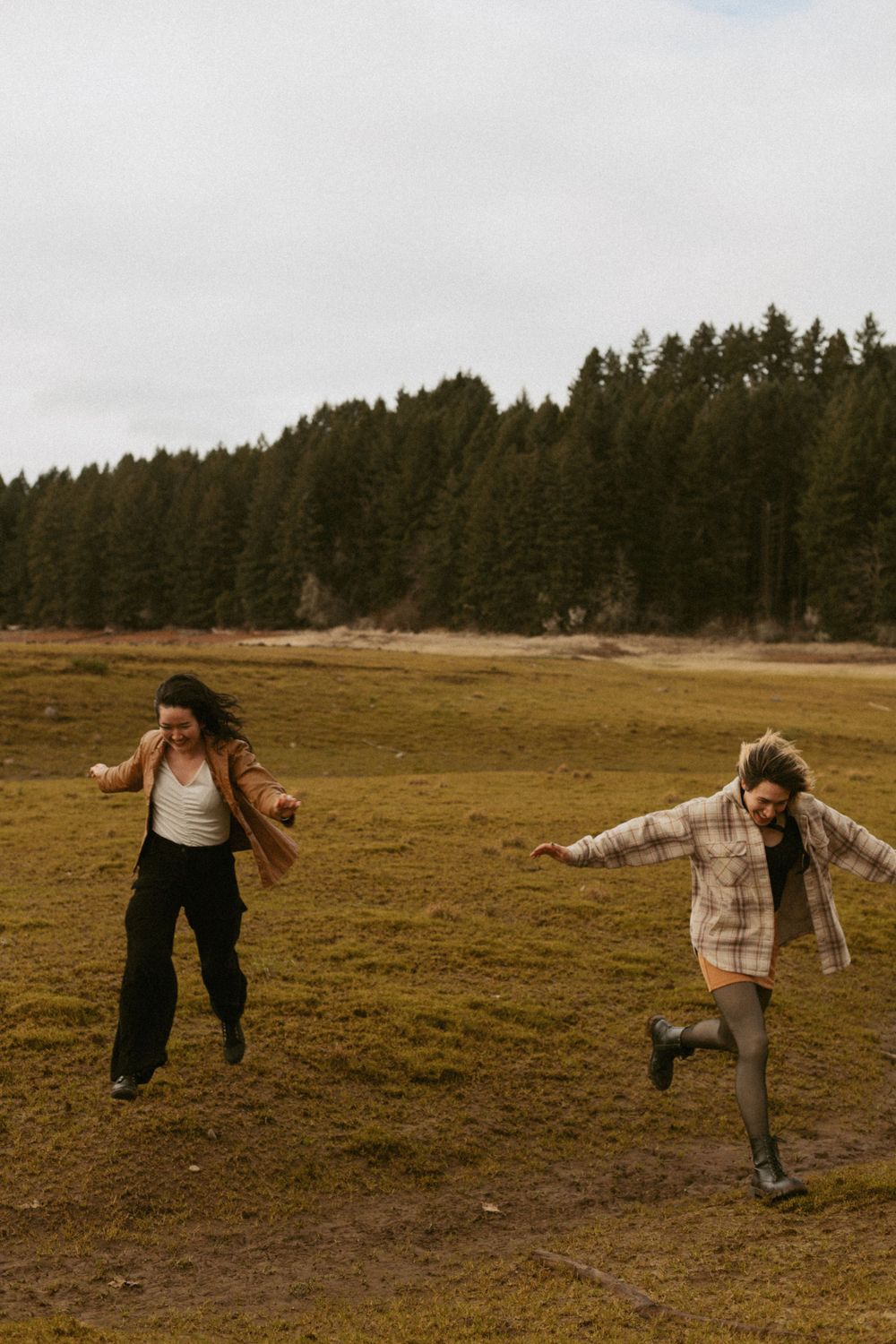 People playfully running and jumping in a field with pine forest backdrop.