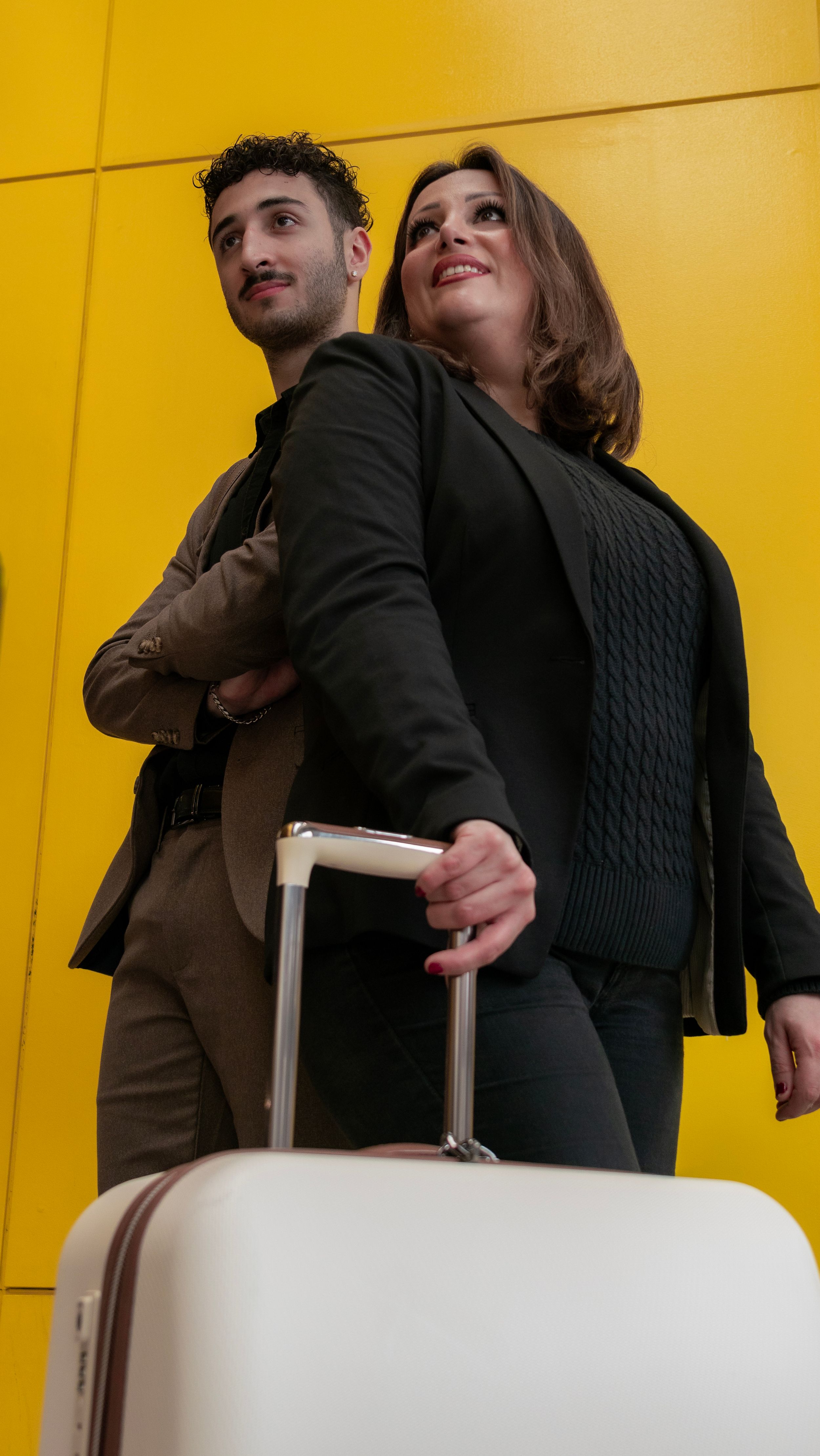 Couple posing playfully with luggage against vibrant yellow wall.