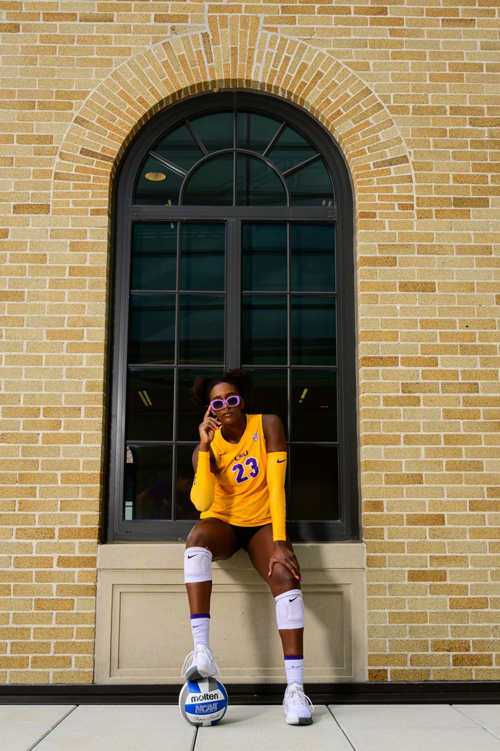 LSU athlete in yellow jersey sits by brick wall with archways during photo shoot.