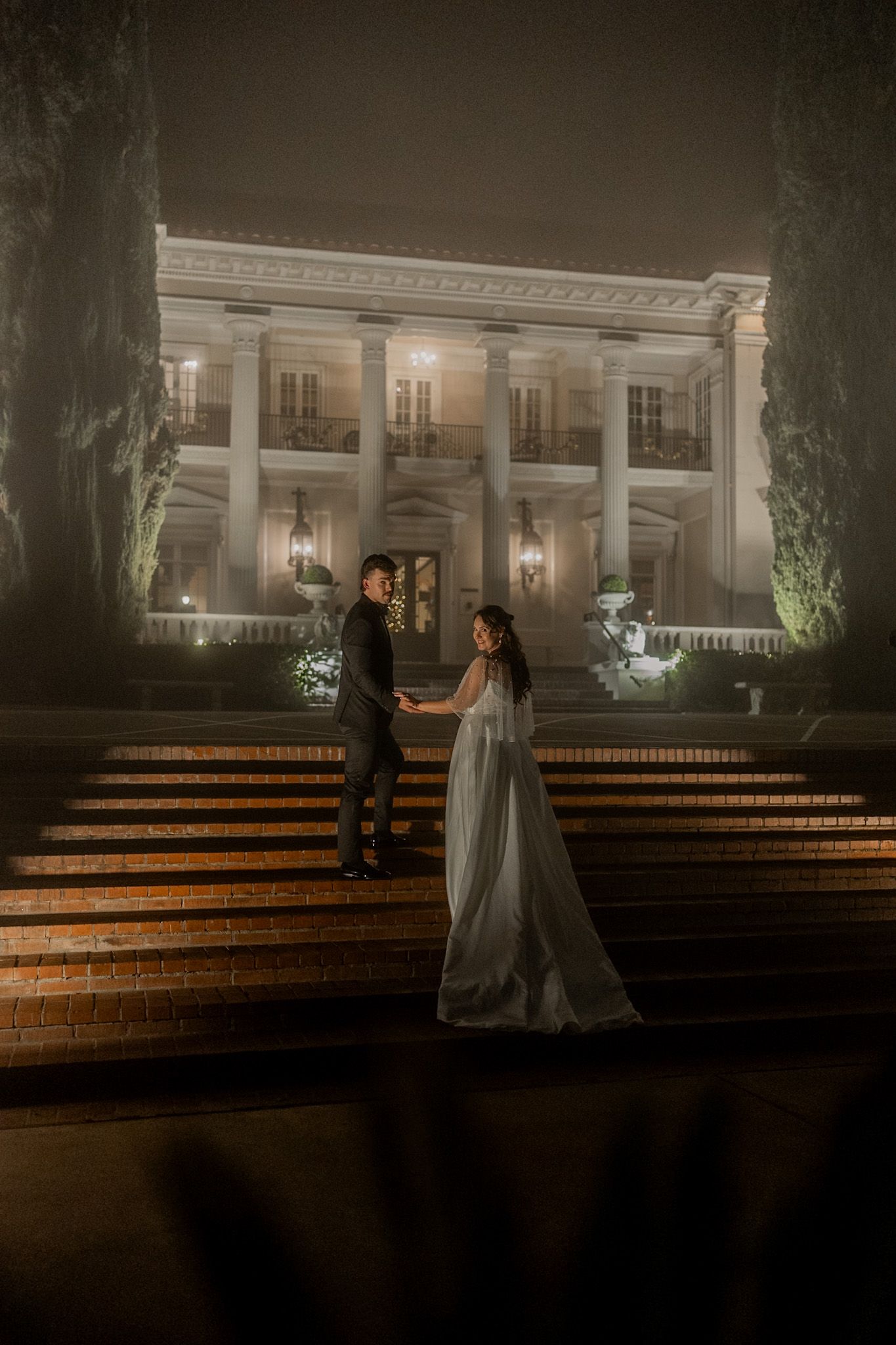 Bride in lace wedding gown poses elegantly among tall pine trees at sunset.