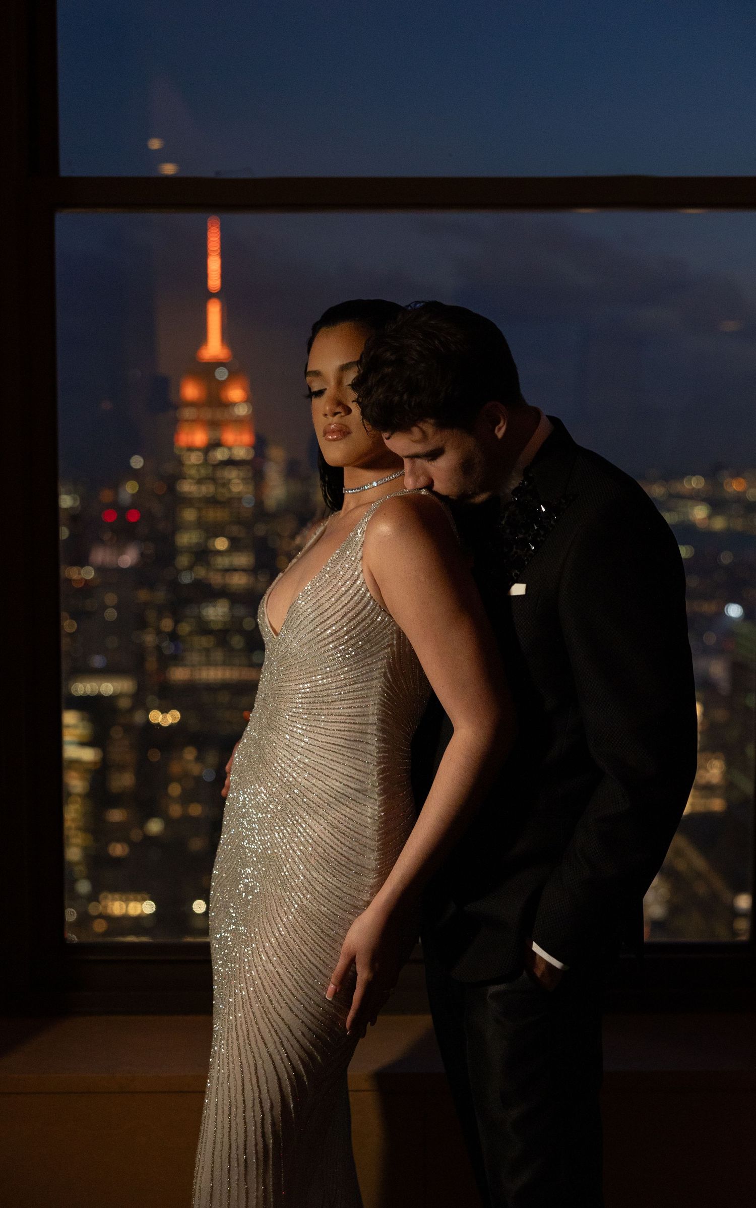 Romantic couple poses against New York City skyline with Empire State Building glowing orange in twilight.