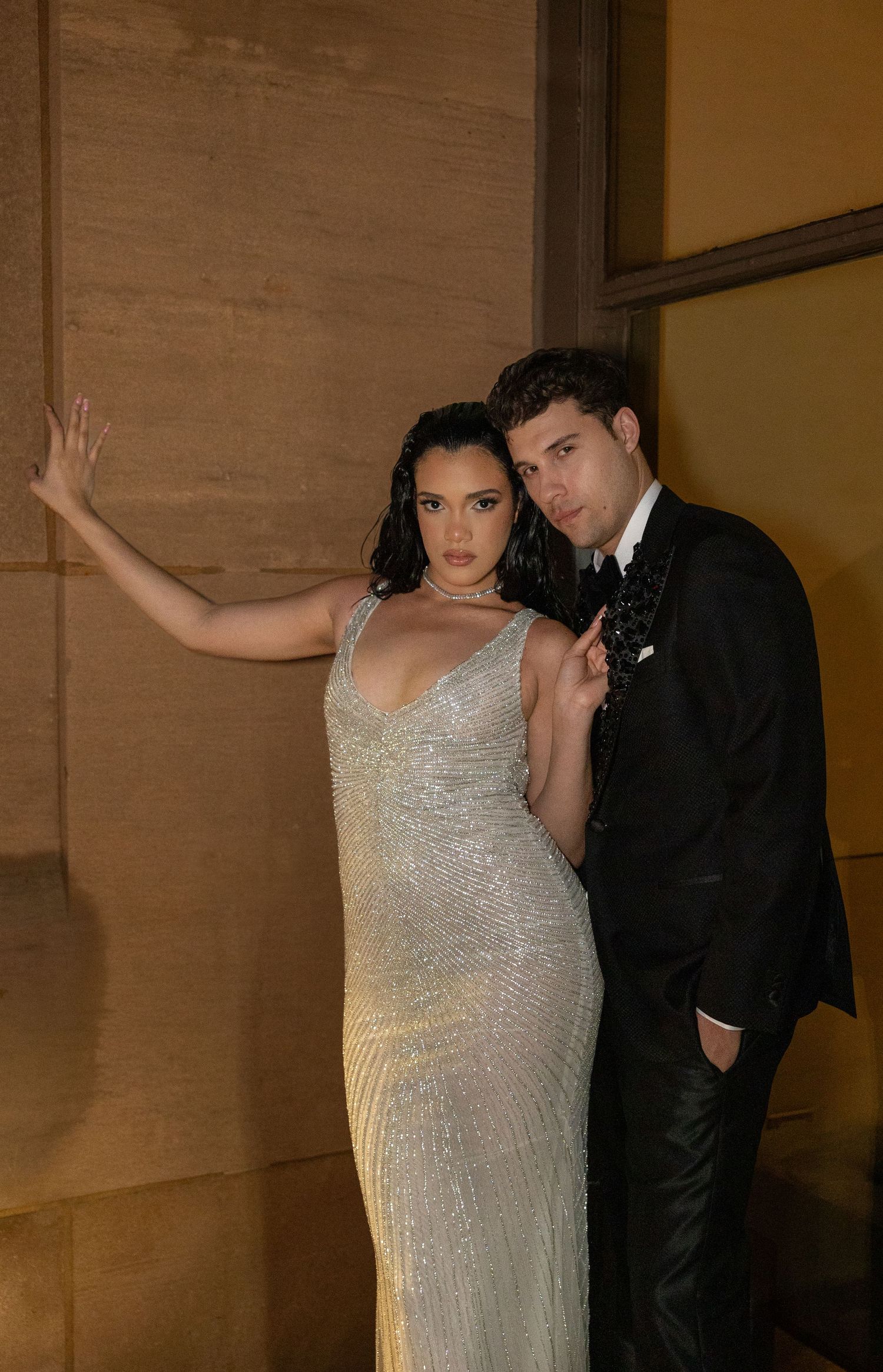 Couple in elegant evening wear poses dramatically against textured wall.
