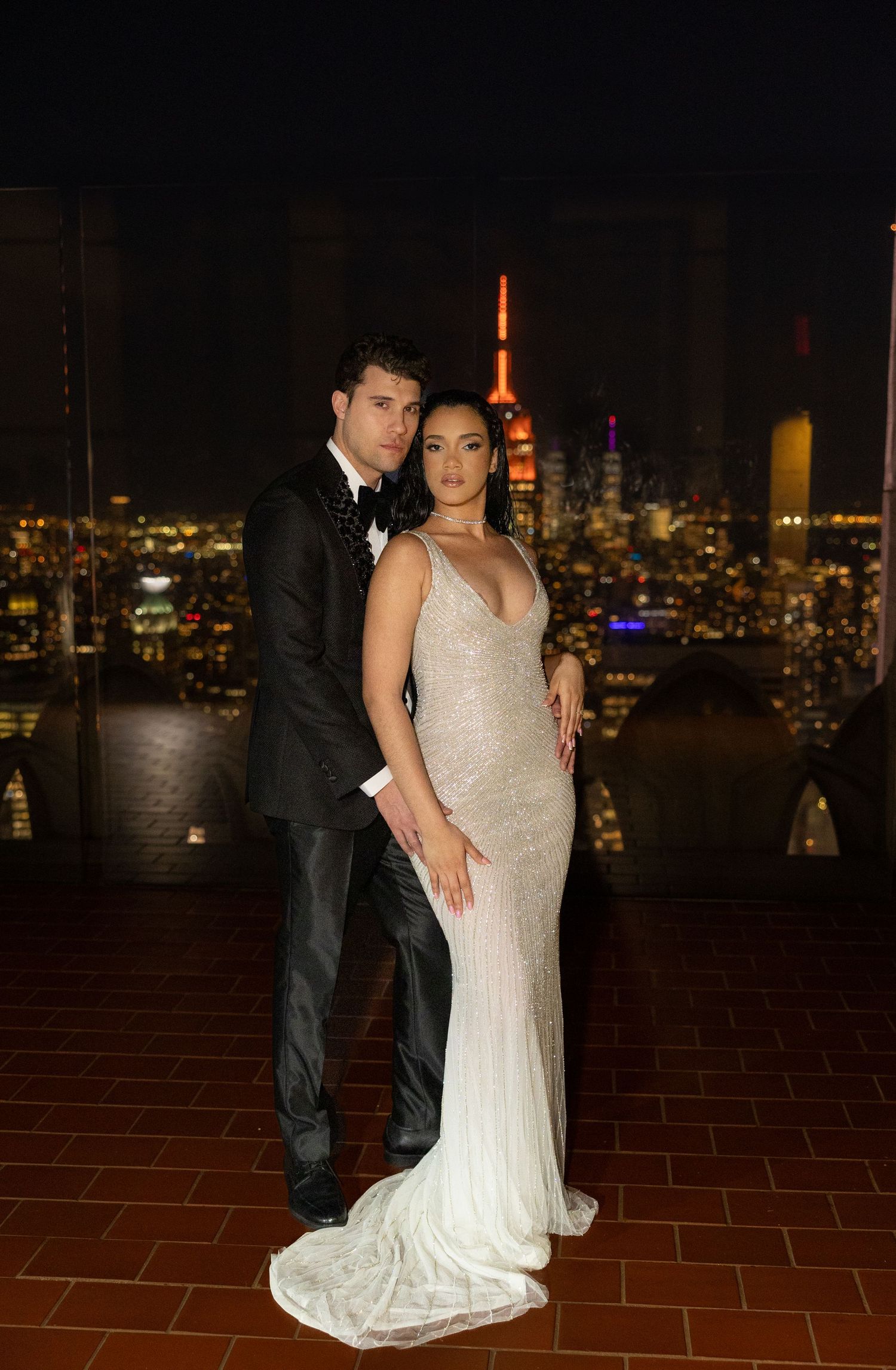 Elegant couple poses on terrace with NYC skyline and Empire State Building glowing orange.