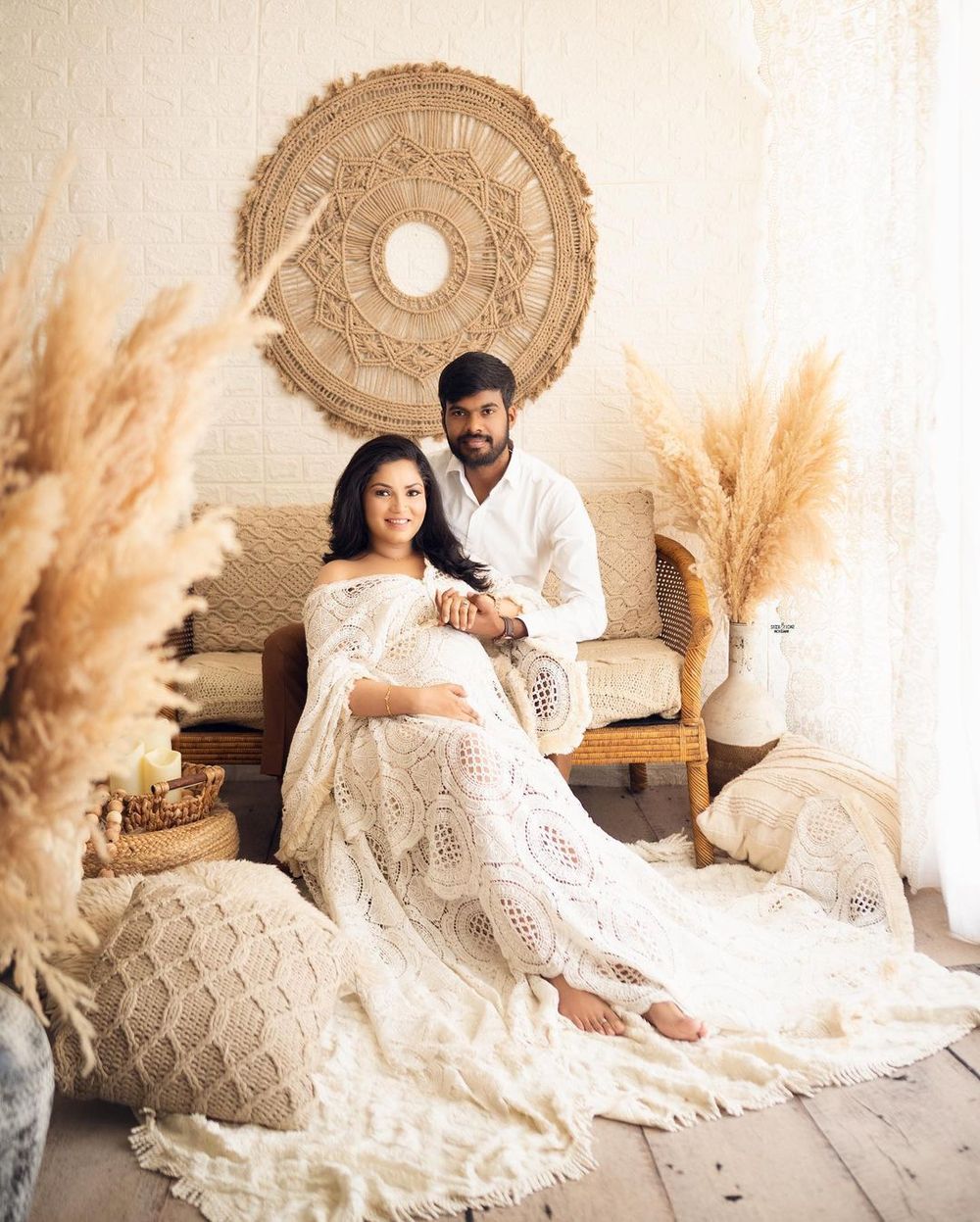 Romantic maternity portrait with couple in white lace sitting on vintage sofa with boho wall decor.