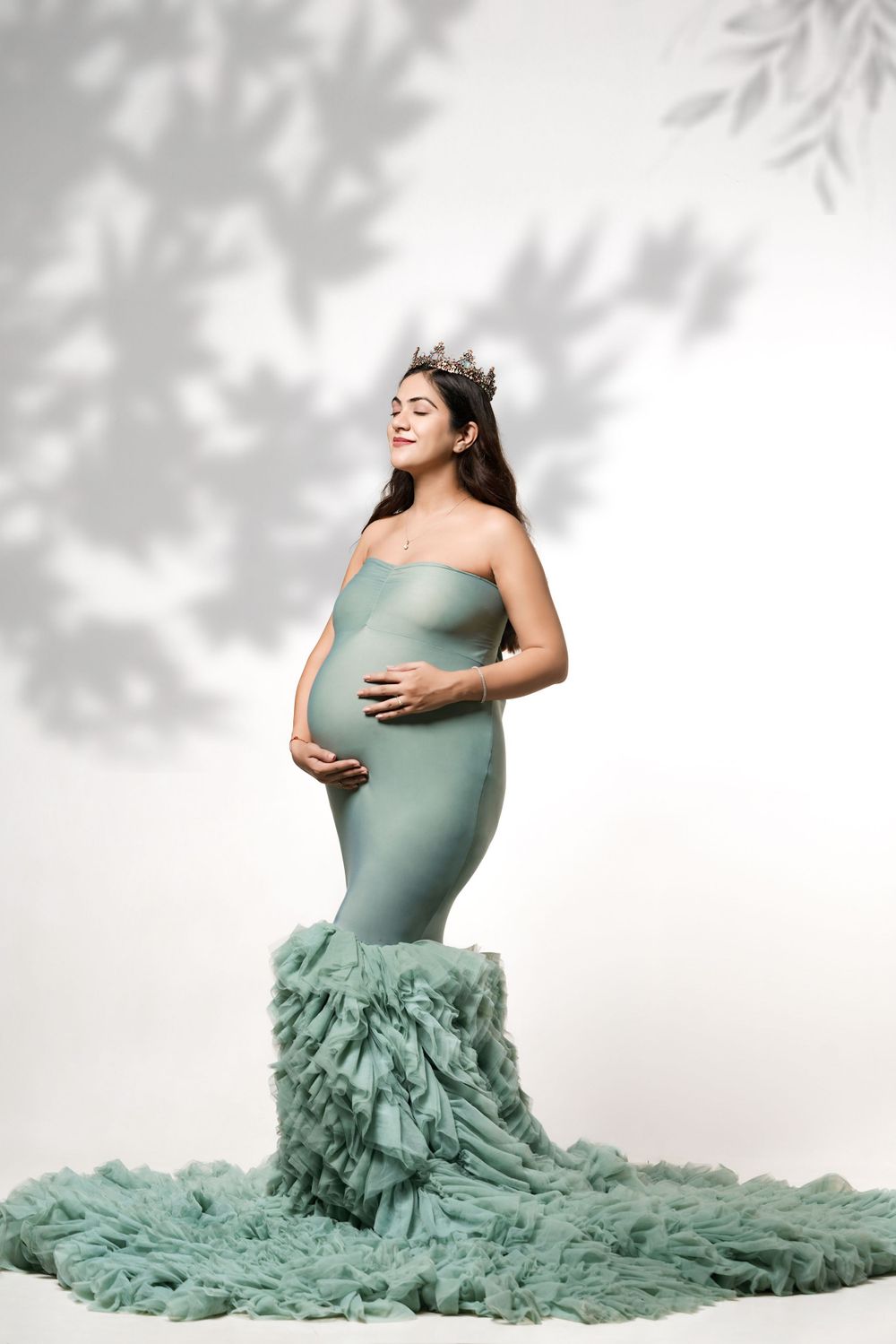 Ethereal maternity portrait in sage green mermaid gown with crown and leaf shadows.