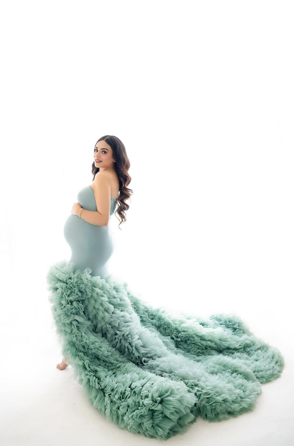 Stunning sage green tulle maternity gown with dramatic ruffled train against white backdrop.