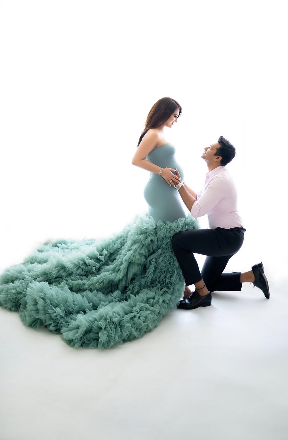 Romantic maternity photo with couple in mint green tulle gown against white backdrop.